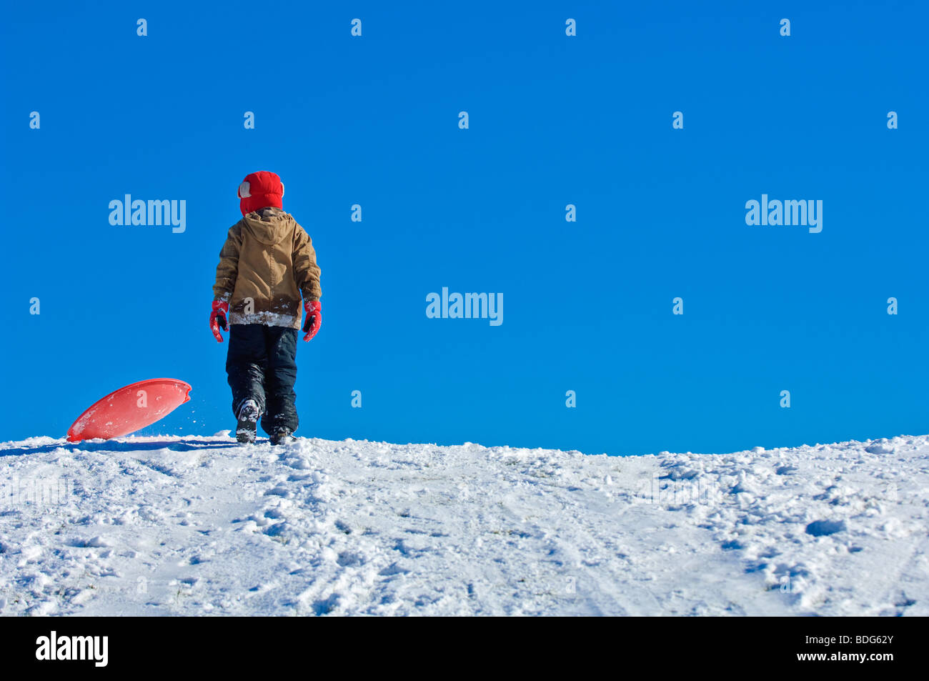 A boy climbs a snowy hill to go sledding under a clear blue sky Stock