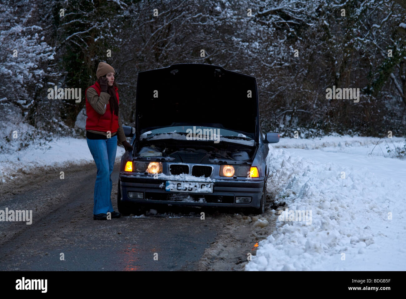 Women on her own broken down in the snow, stranded trying to get it fixed on mobile phone ringing rescue service at night. Stock Photo