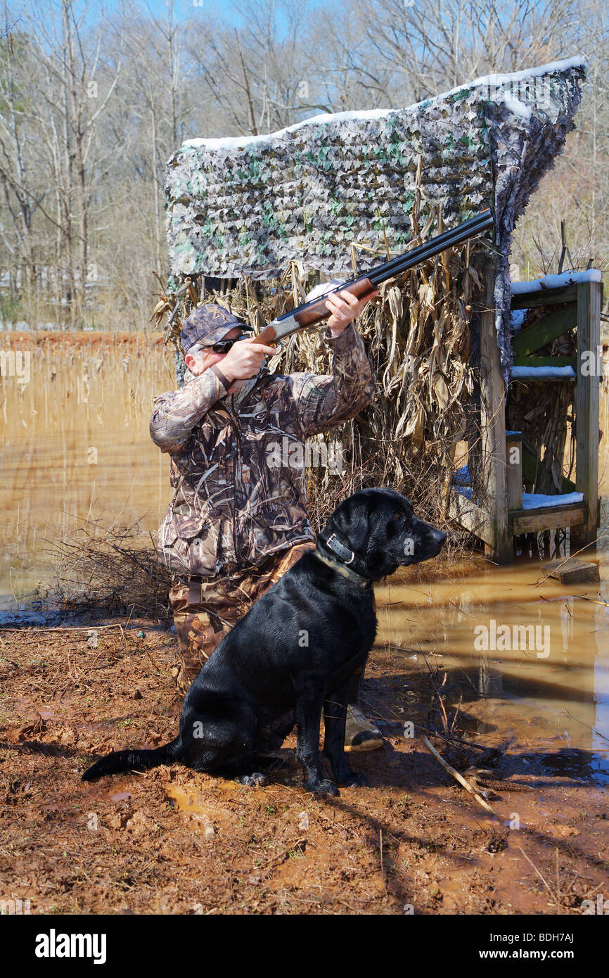 DUCK HUNTER IN CAMO AND BLACK LAB LABRADOR ON SHORE WAITING FOR WATERFOWL MALLARDS BROWNING CITORI SHOTGUN SNOW ON GROUND BLIND Stock Photo