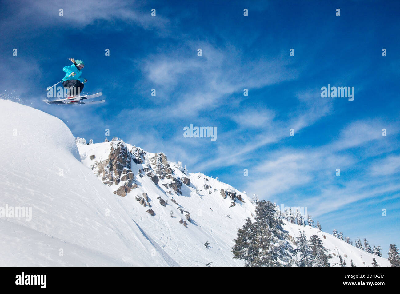 A female skier launching into fresh powder snow with mountains and blue sky in the background. Stock Photo