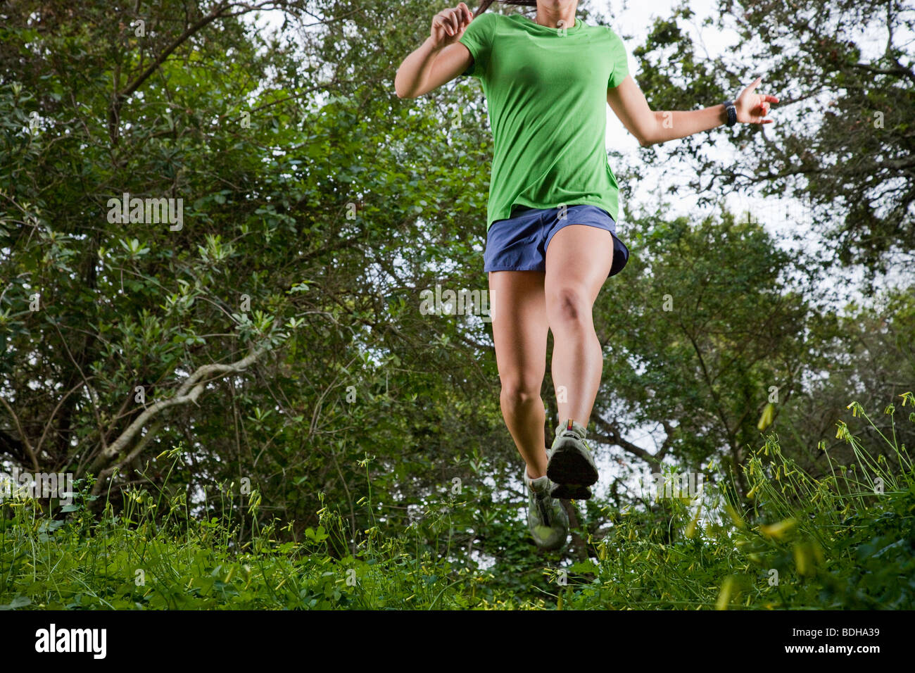 View from below of a woman trail running through a green meadow in the woods. Stock Photo