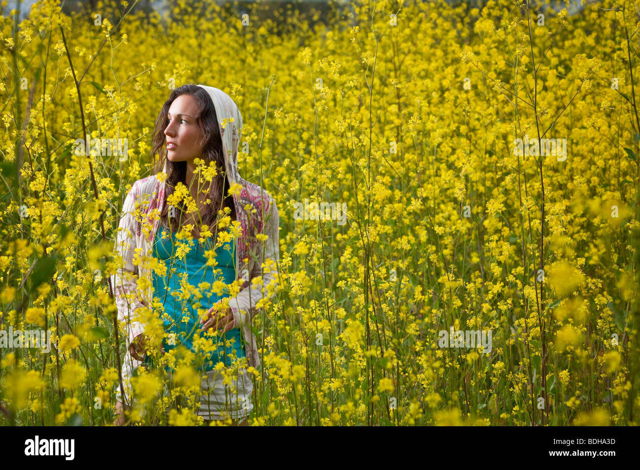 Young woman standing in a field of yellow flowers wearing a hood and looking into the distance Stock Photo