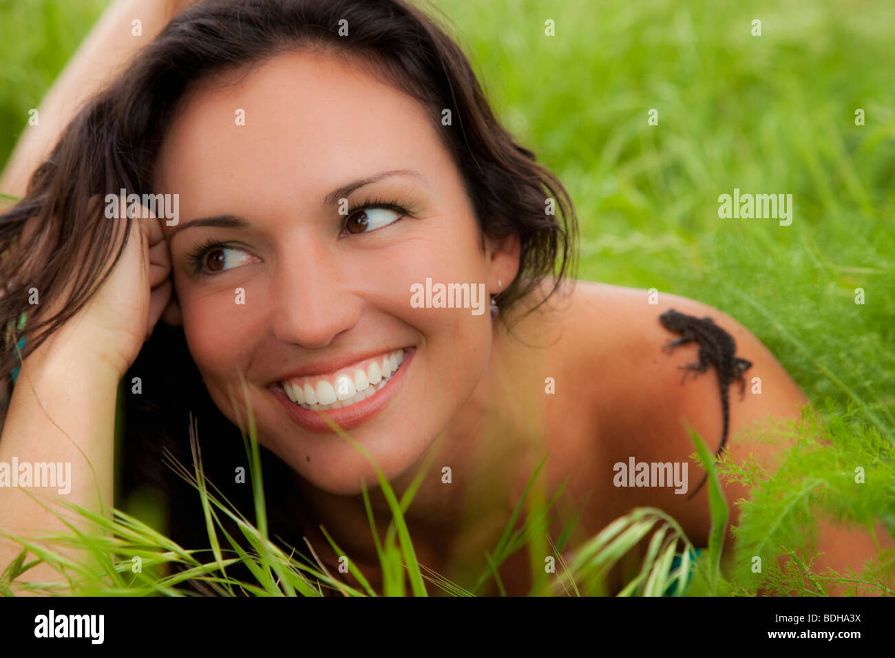 Happy young woman in a field of fresh grass smiles as a small lizard crawls up her shoulder. Stock Photo