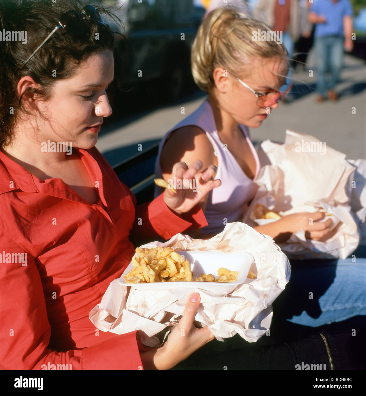 Two young women sitting on a bench eating fish and chips Tenby Pembrokeshire South Wales Stock Photo