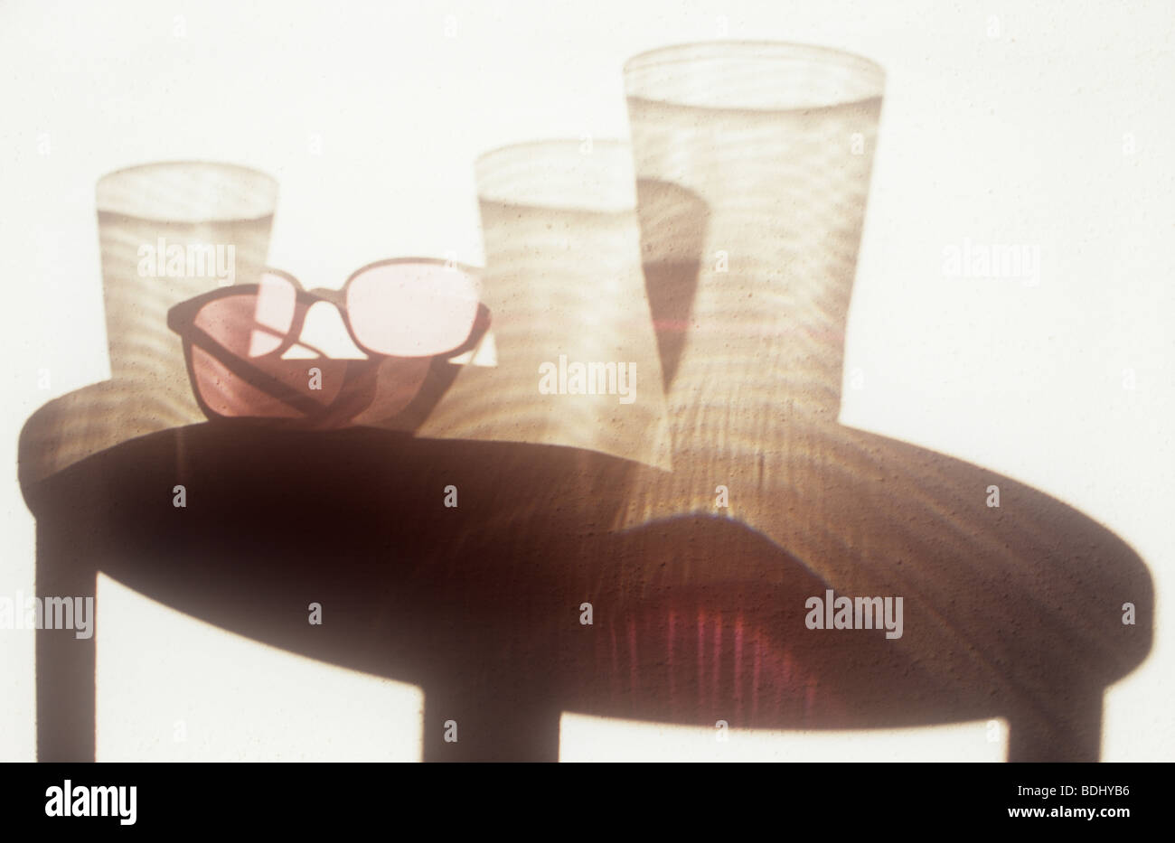 Shadow on white wall of circular table with three empty glasses and rose-tinted spectacles casting refracted reflections Stock Photo