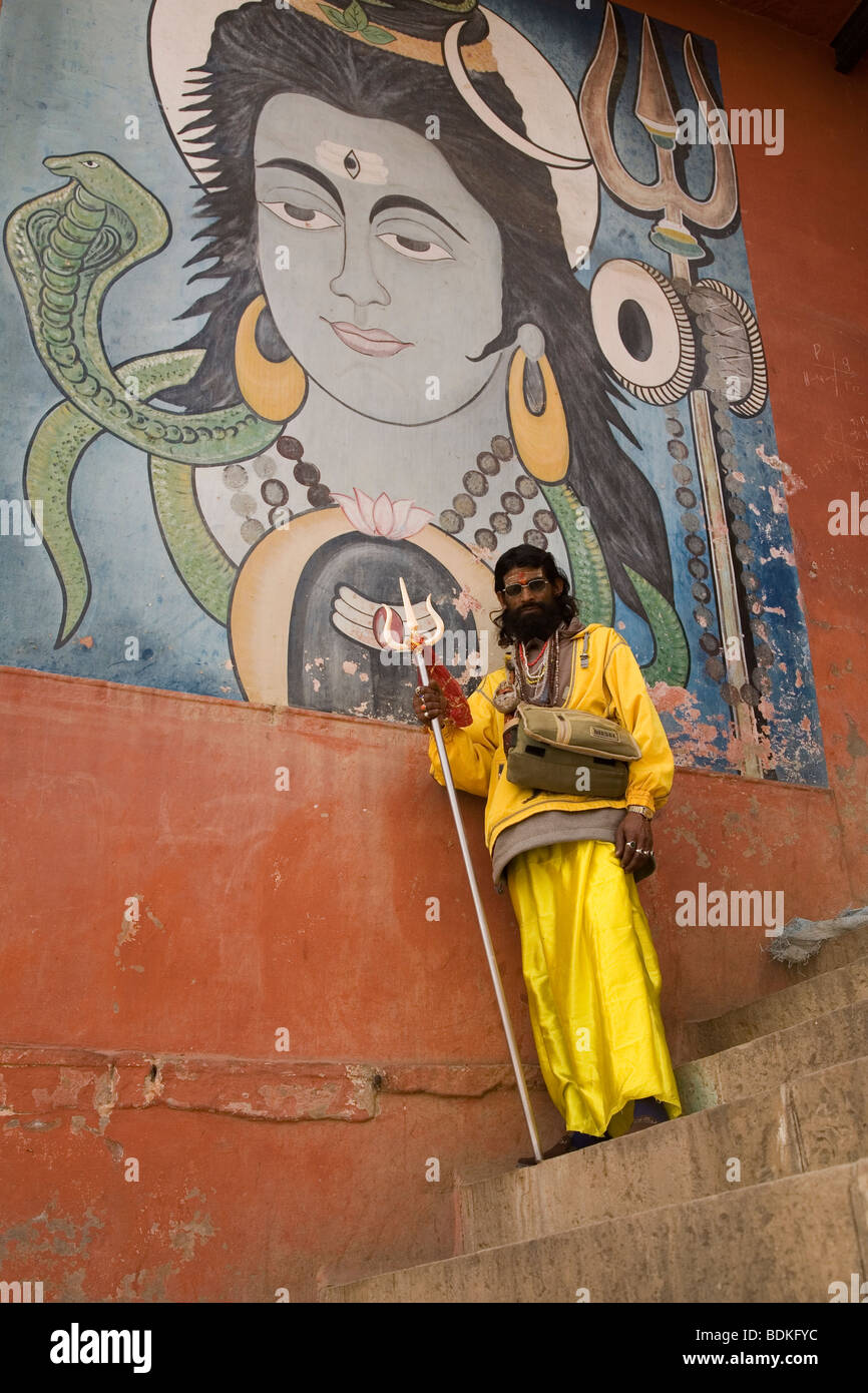 A Sadhu in the Indian city of Varanasi (Benares). He stands in front of a painted image of the Hindu god Shiva. Stock Photo