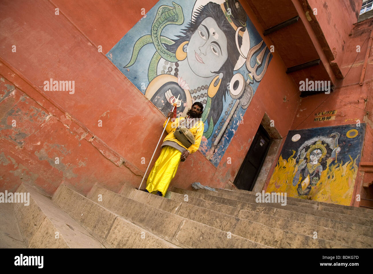 A Sadhu in the Indian city of Varanasi (Benares). He stands in front of a painted image of the Hindu god Shiva. Stock Photo