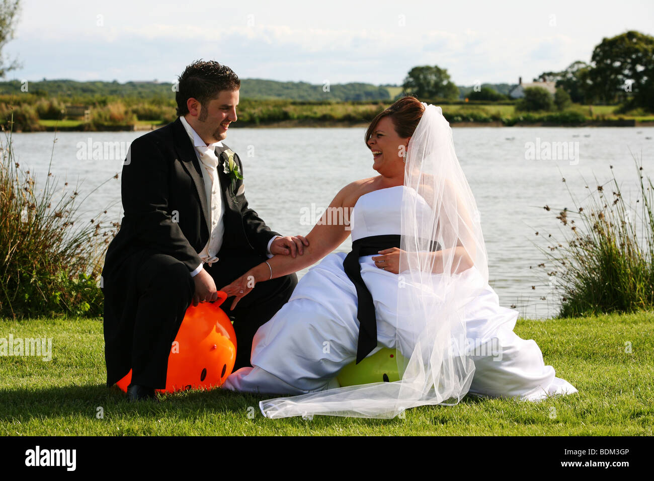 Bride and Groom relax have fun on colourful space hopper toys on wedding day next to edge of lake on grass UK Stock Photo