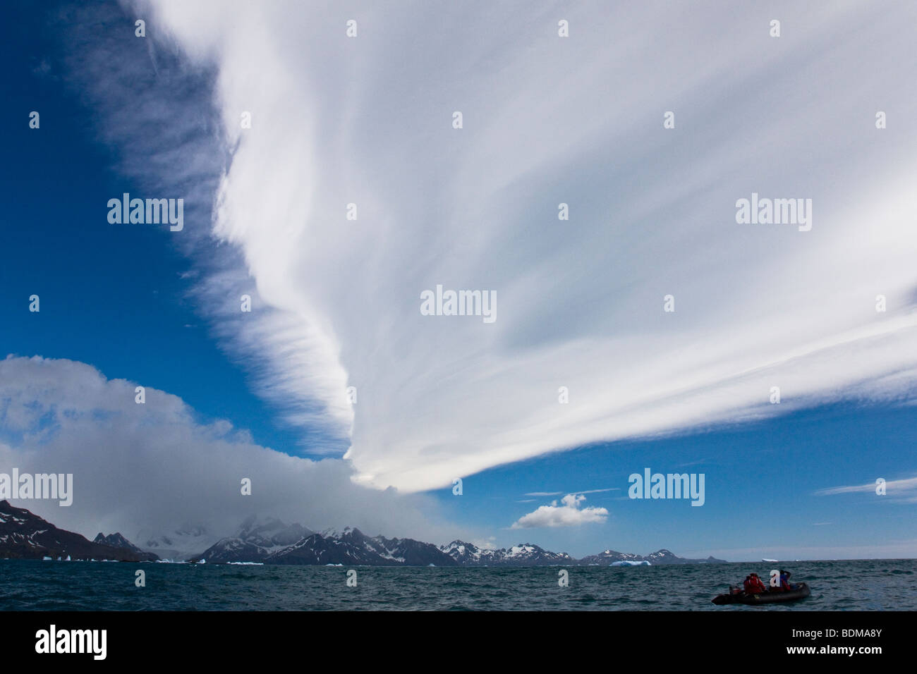Tall altocumulus lenticular clouds form above tall mountain in Antarctic summer, South Georgia Antarctica small zodiac boat with people in foreground Stock Photo