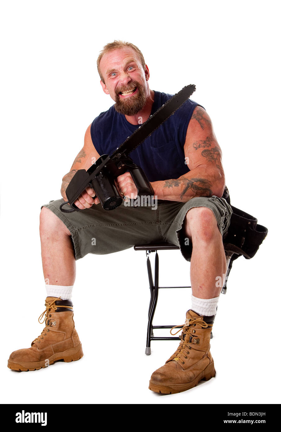 Crazy Caucasian man with tattoos and chainsaw sitting on stool with strong facial expression, isolated. Stock Photo