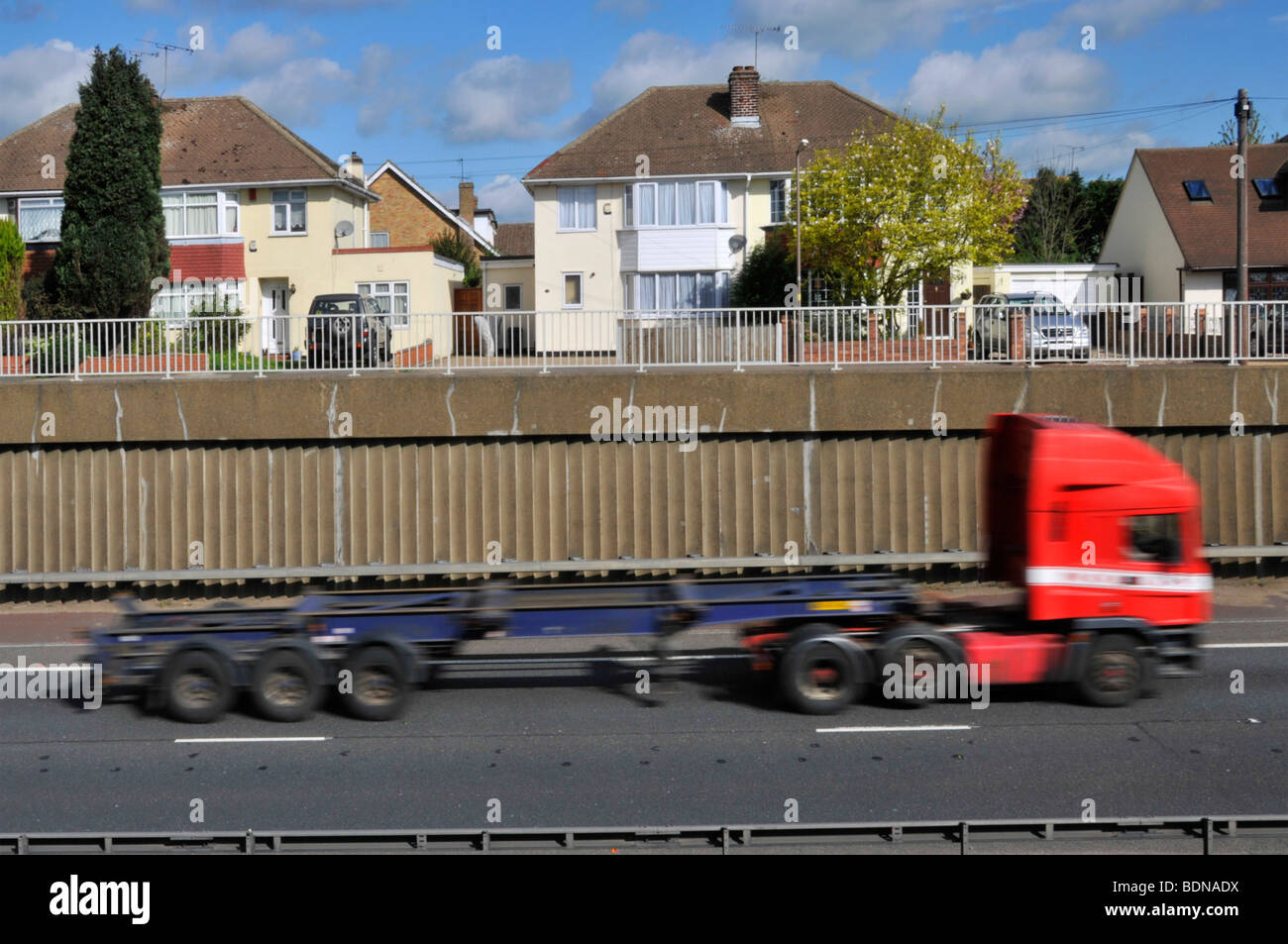 Blurred fast articulated trailer and lorry in cutting on A12 dual carriageway passing semi detached houses Stock Photo