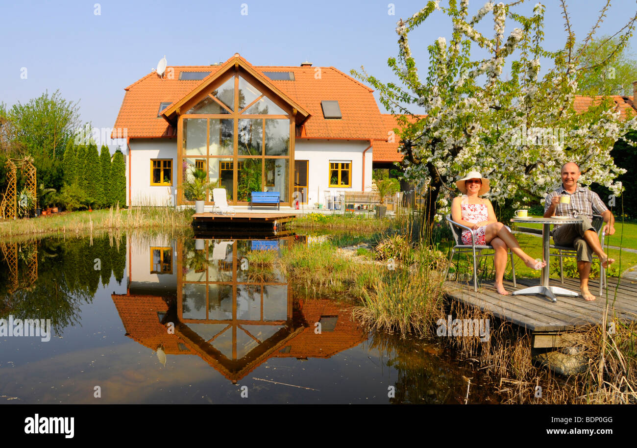Couple drinking coffee at a garden pond in front of a house with conservatory and flowering cherry tree Stock Photo