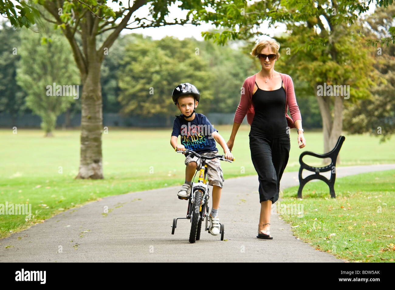 Horizontal close up portrait of a young mum helping her son learn to ride a bike with stabilisers in the park Stock Photo