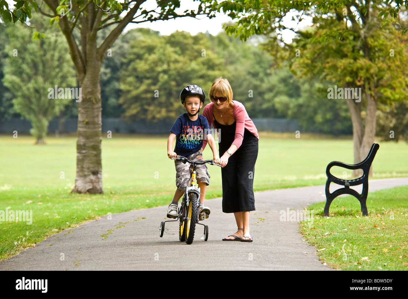 Horizontal close up portrait of a young mum helping her son learn to ride a bike with stabilisers in the park Stock Photo
