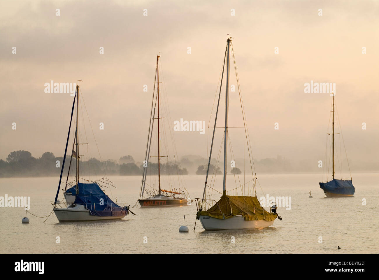 The first light of the day hits sailing boats in front of thick morning fog at Ammersee lake near Schondorf, Bavaria, Germany,  Stock Photo