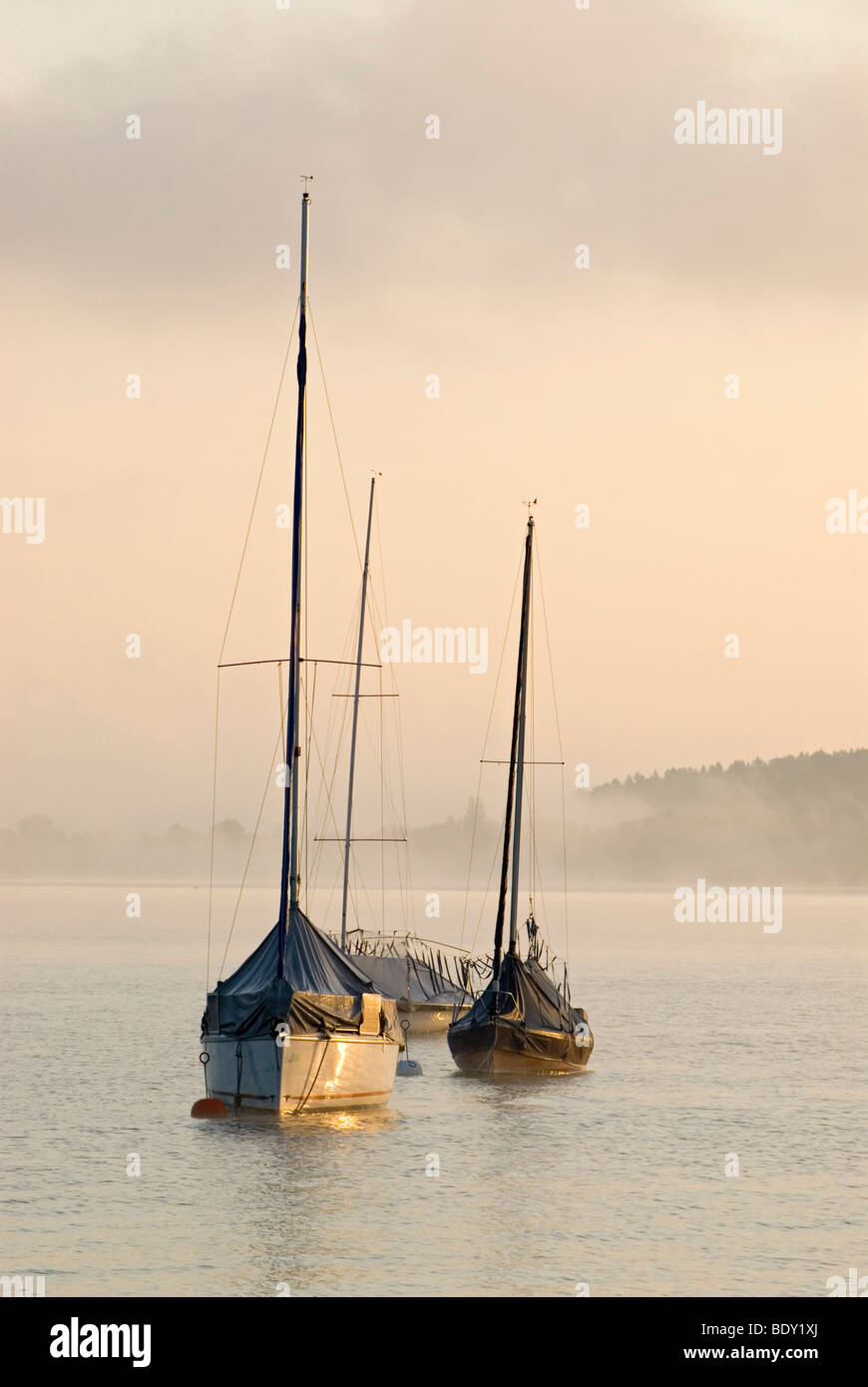 The first light of the day hits sailing boats in front of thick morning fog at Ammersee lake near Schondorf, Bavaria, Germany,  Stock Photo