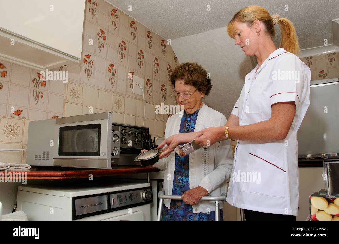 Elderly woman in kitchen learns to cook meal for one in a microwave oven and being suppervised by a carer Stock Photo