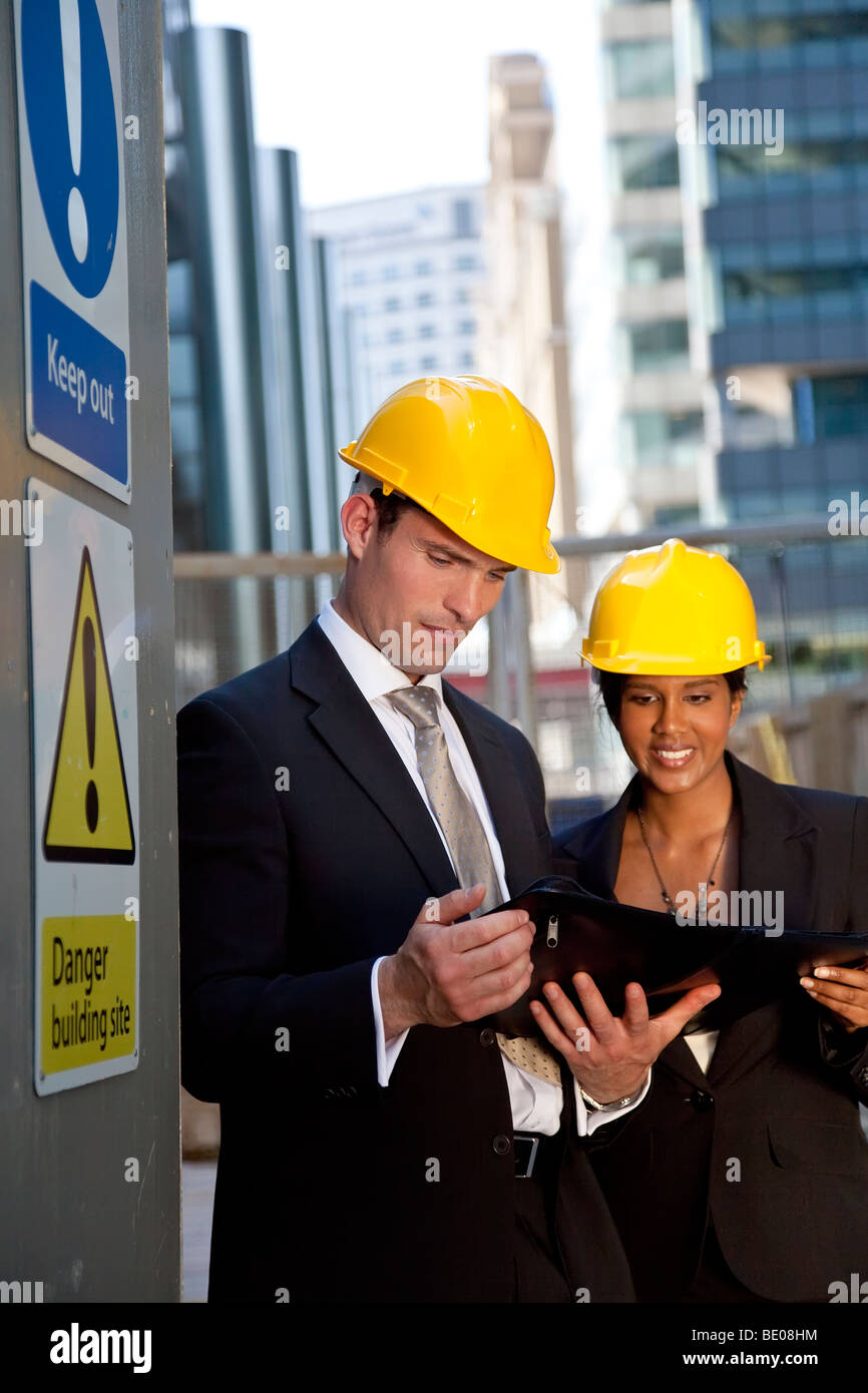 A male manager on a construction site wearing a hard hat and talking with his Asian female colleague Stock Photo