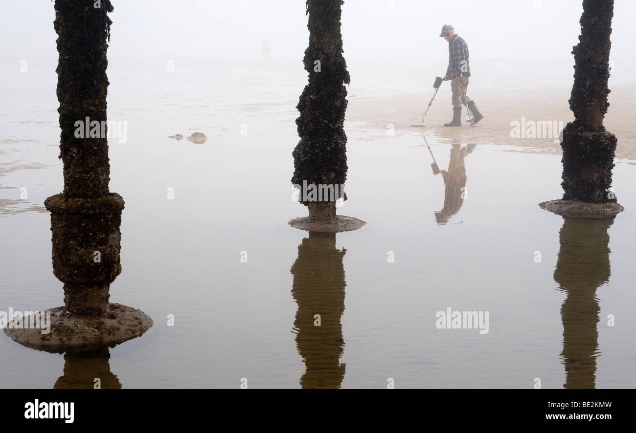 A metal detector searches the beach and sand on a misty day under the legs of a victiorian pier on the east coast of England. Stock Photo