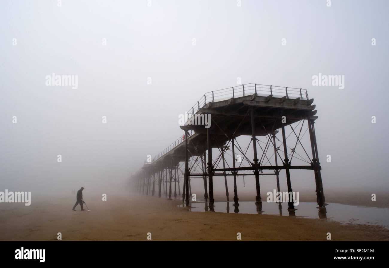 A metal detector searches the beach and sand on a misty day under the legs of a victiorian pier on the east coast of England. Stock Photo