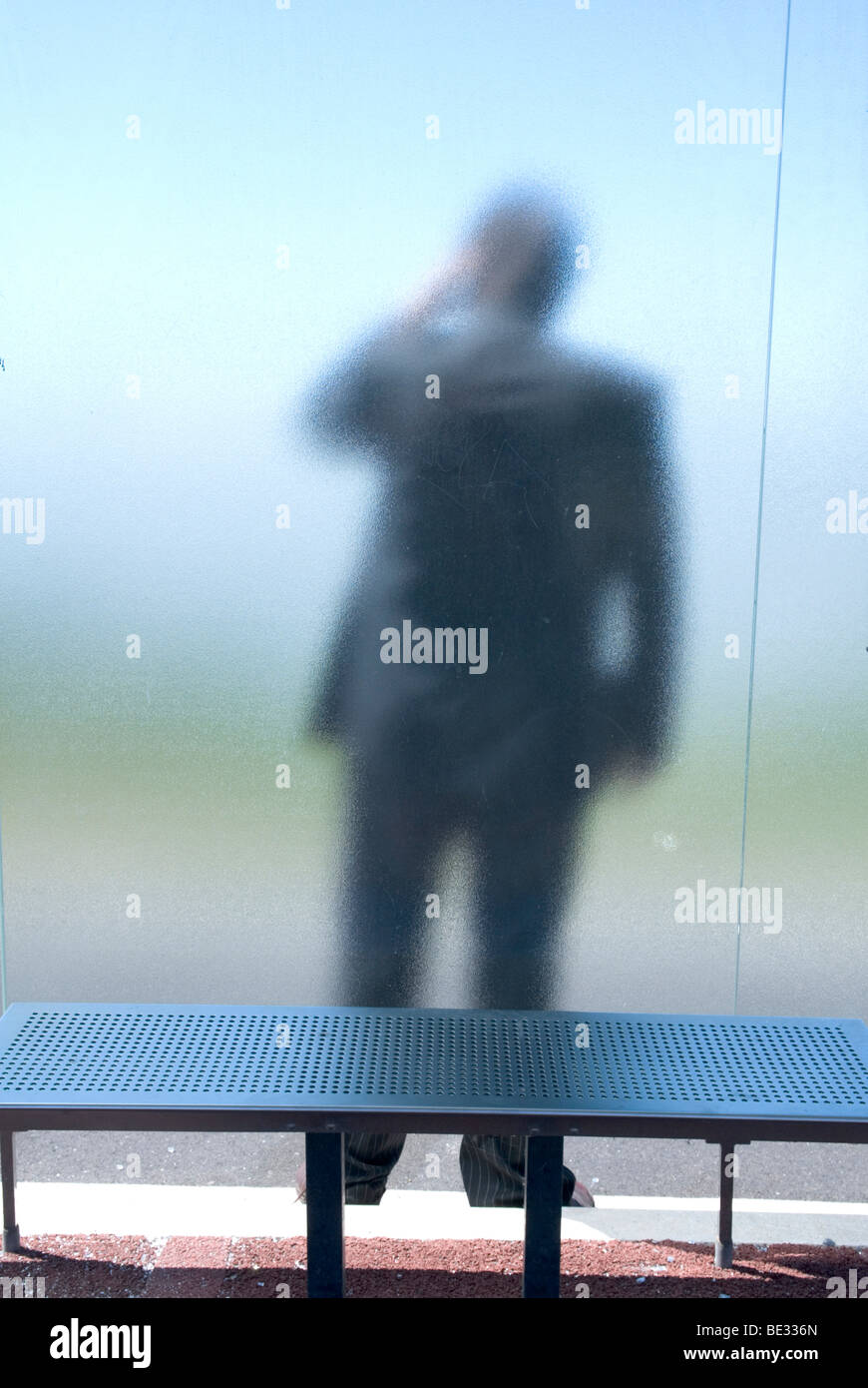 a man in a suit, with his back to us, stands behind frosted glass at a bus stop Stock Photo