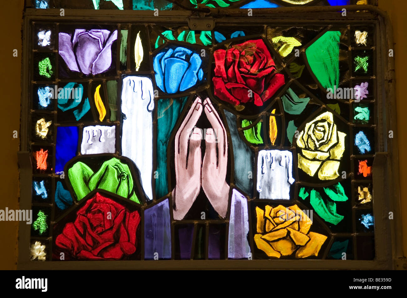 Stained window with clasped hands in sign of prayer, candles and colorful flowers Stock Photo