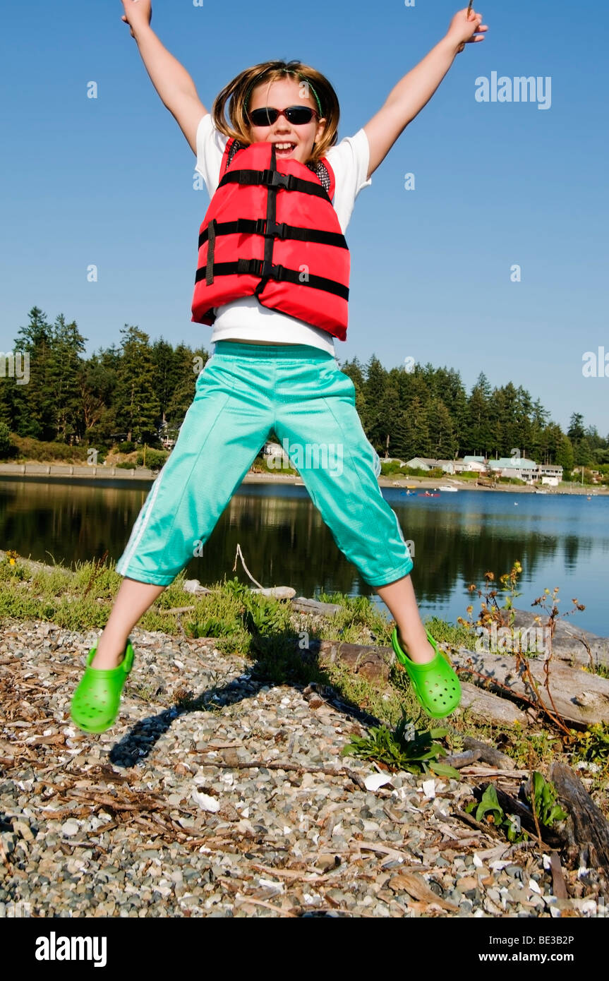 A cute little girl wearing a life jacket jumps for joy while playing near the water. Stock Photo