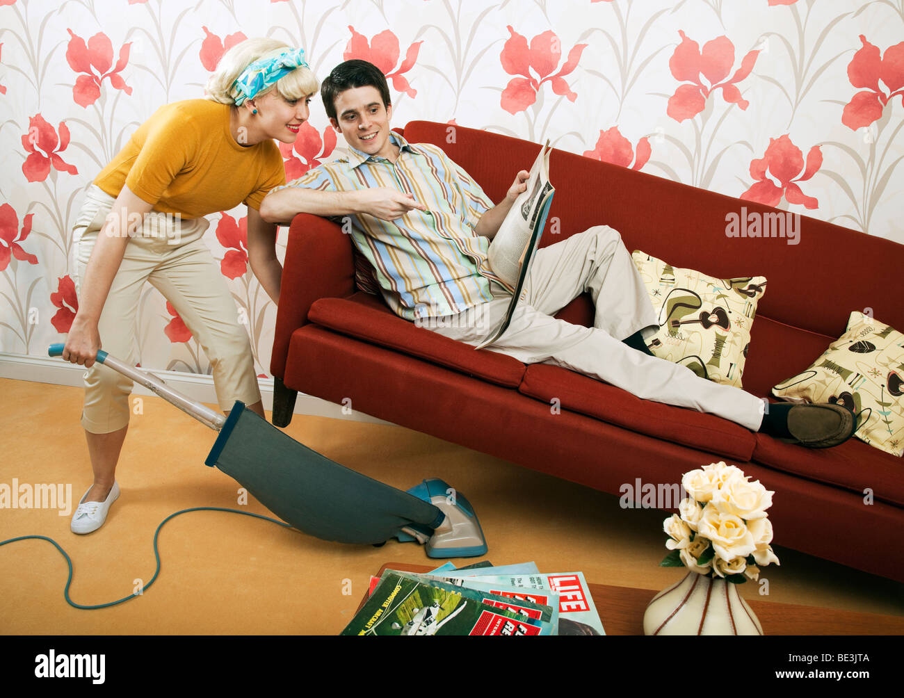 Retro 1960s couple, man on sofa, woman vacuuming Stock Photo