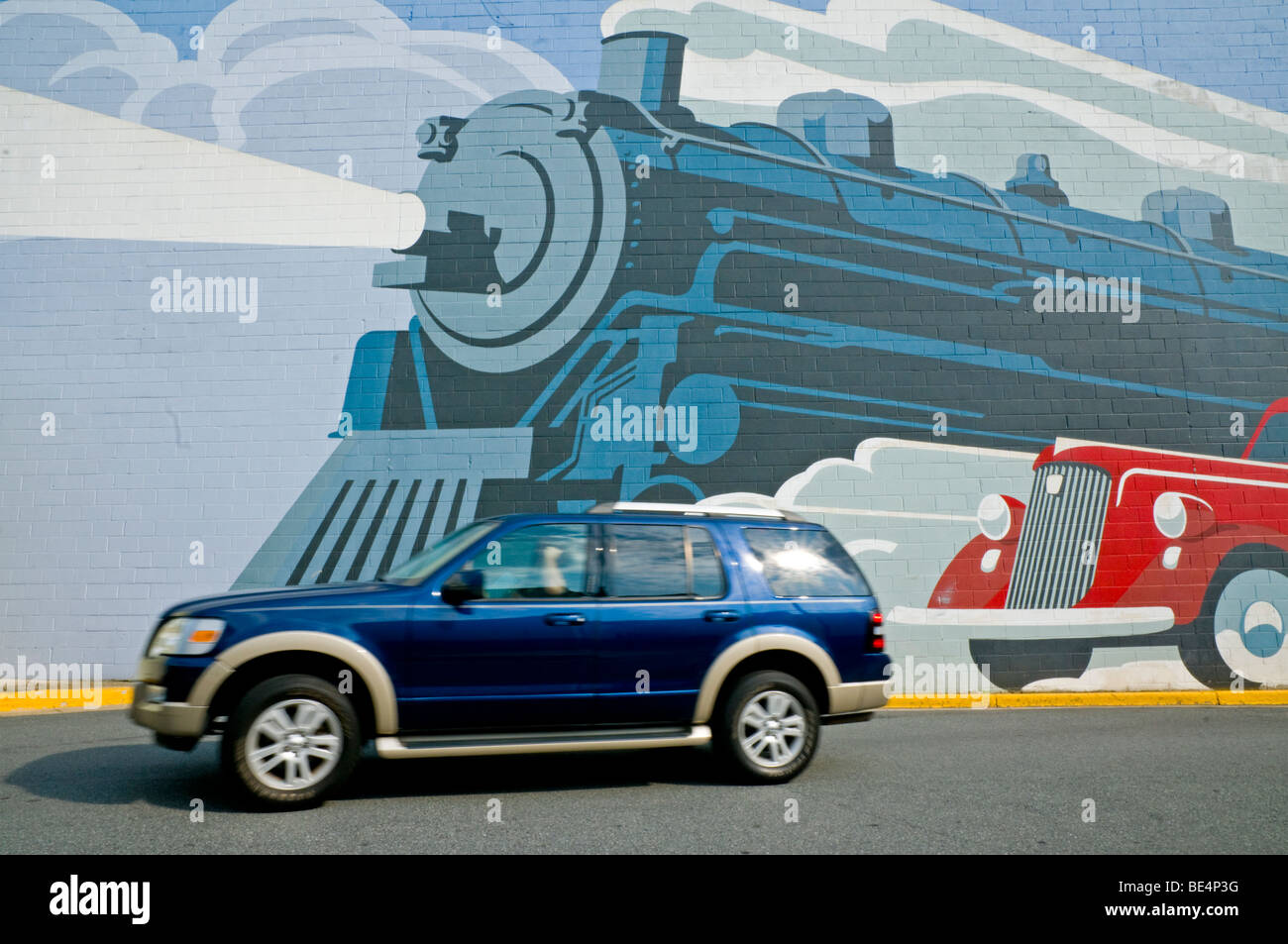 A blue Ford SUV passing in front of a mural featuring a vintage train and automobile. Stock Photo