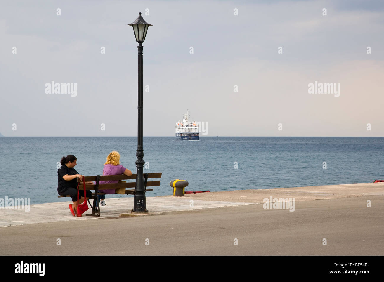 Have they missed the boat. 2 women sitting on bench with ship sailing away in distance Stock Photo