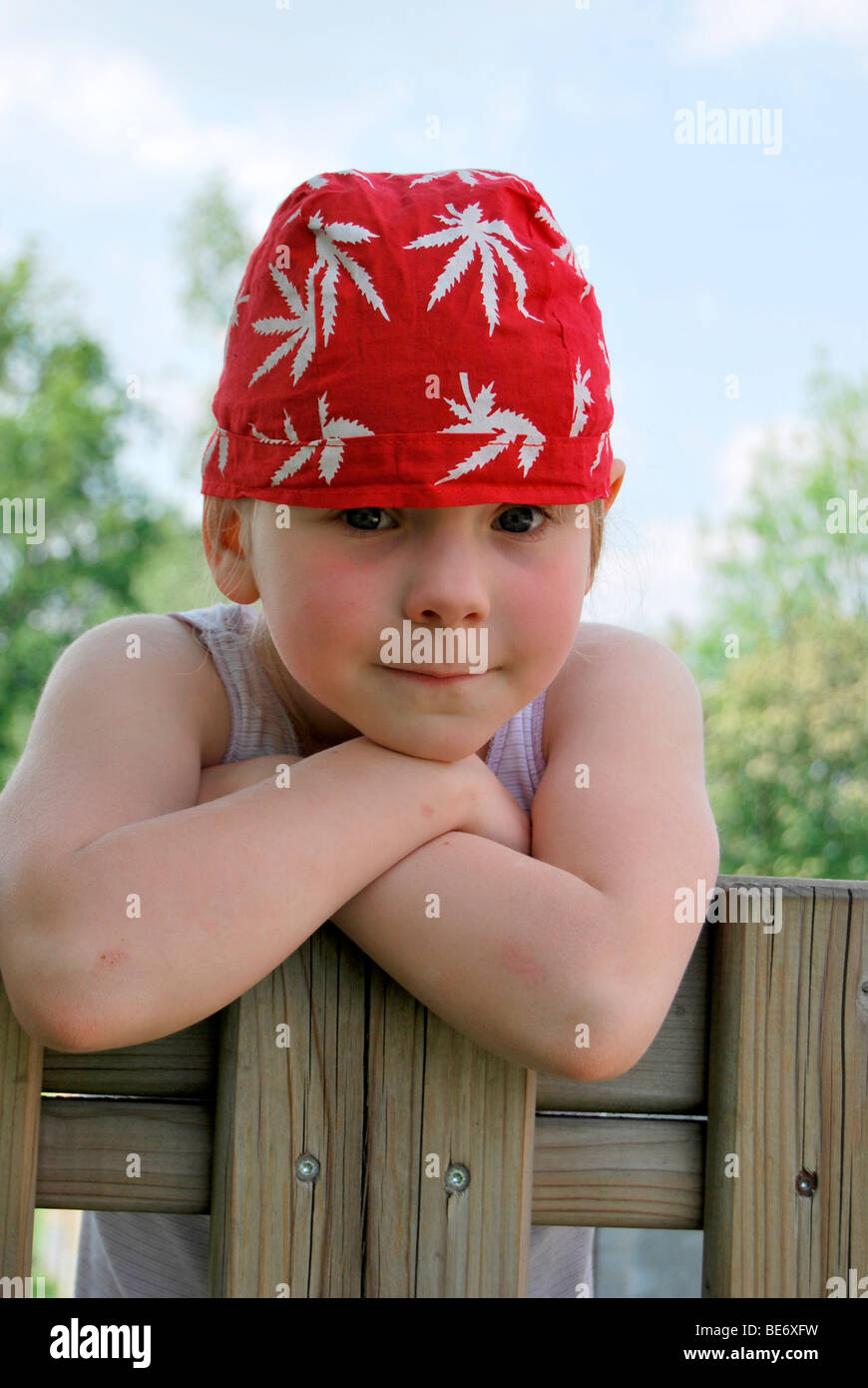 Girl, 5, wearing a red headscarf looking interested while hanging over a garden fence like a young rascal Stock Photo