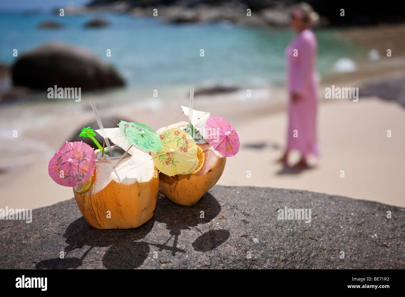 Two decorated coconuts filled with drinks standing on a granite rock, in the back a woman in a pink tunic, island Mahe, Seychel Stock Photo