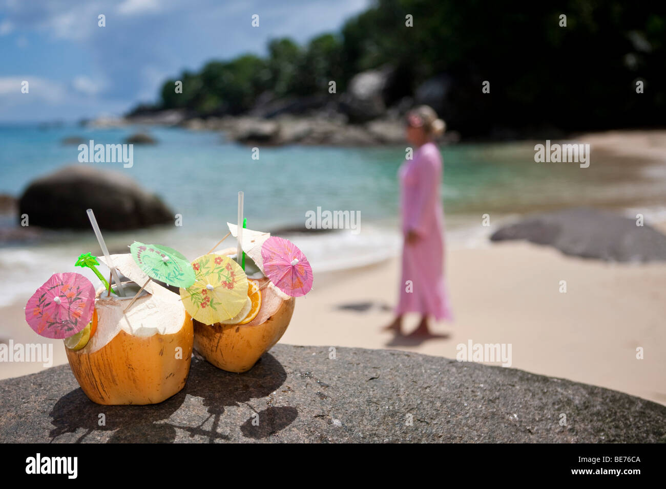 Two decorated coconuts filled with drinks standing on a granite rock, in the back a woman in a pink tunic, island Mahe, Seychel Stock Photo