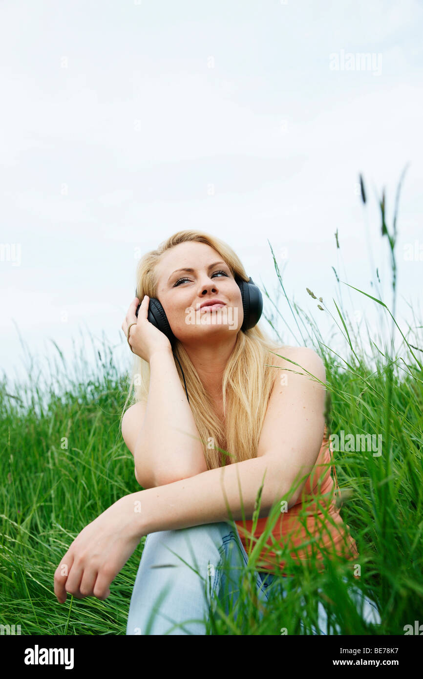 Young blonde woman sitting on a lawn, listening to music with headphones Stock Photo