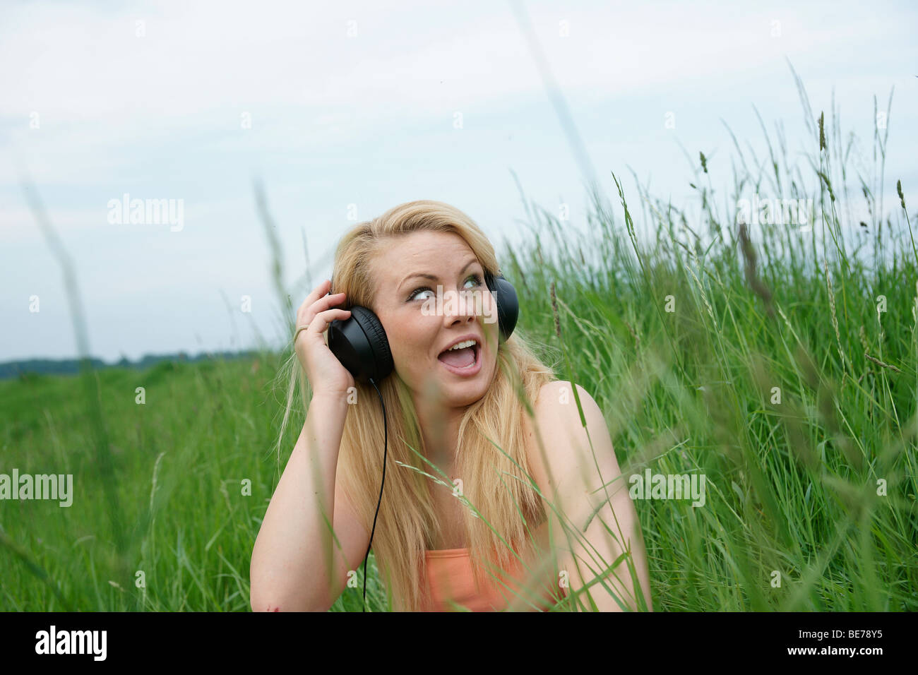 Young blonde woman sitting on a lawn, listening to music with headphones Stock Photo