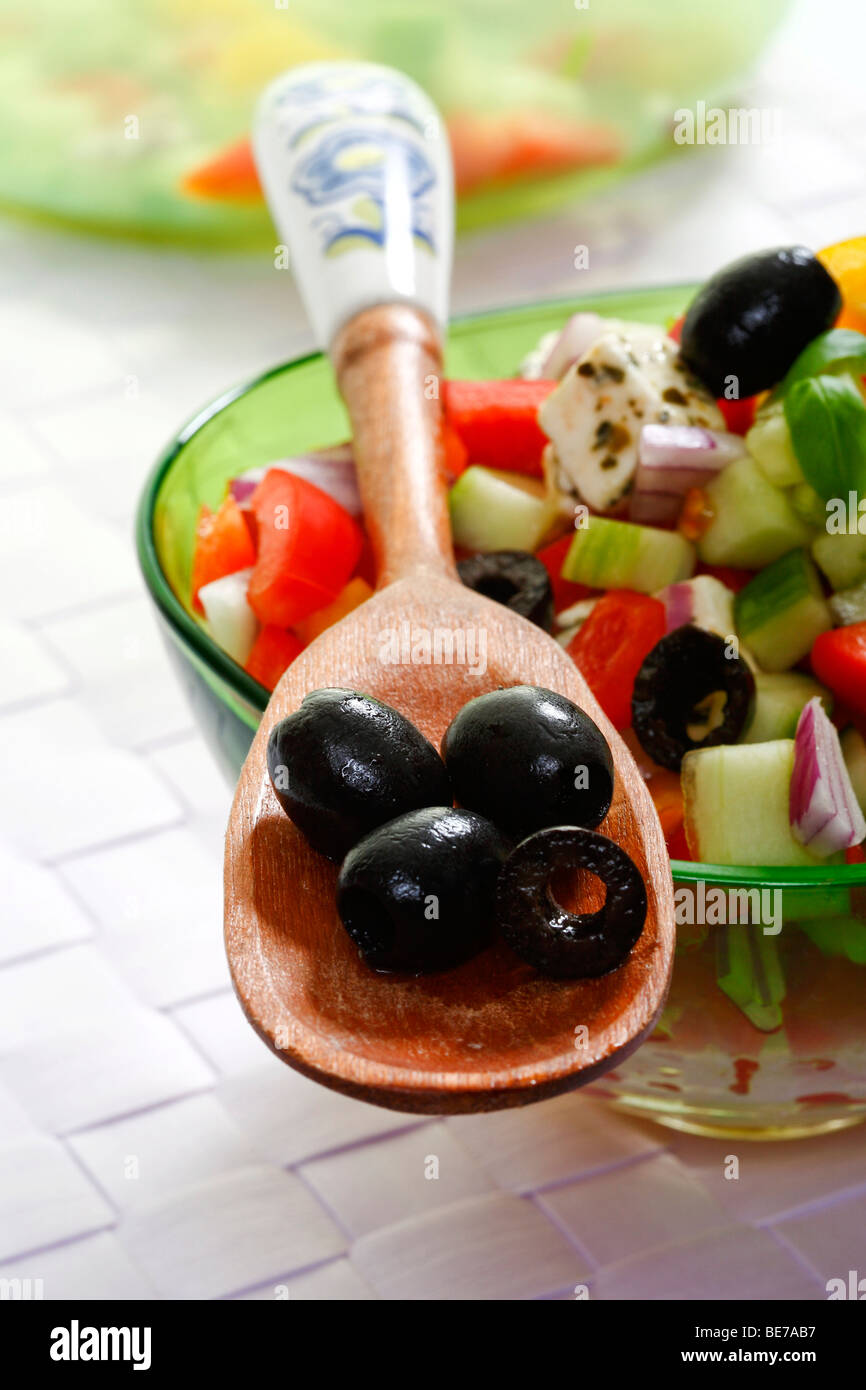 Black olives on a wooden spoon in front of a Greek salad Stock Photo
