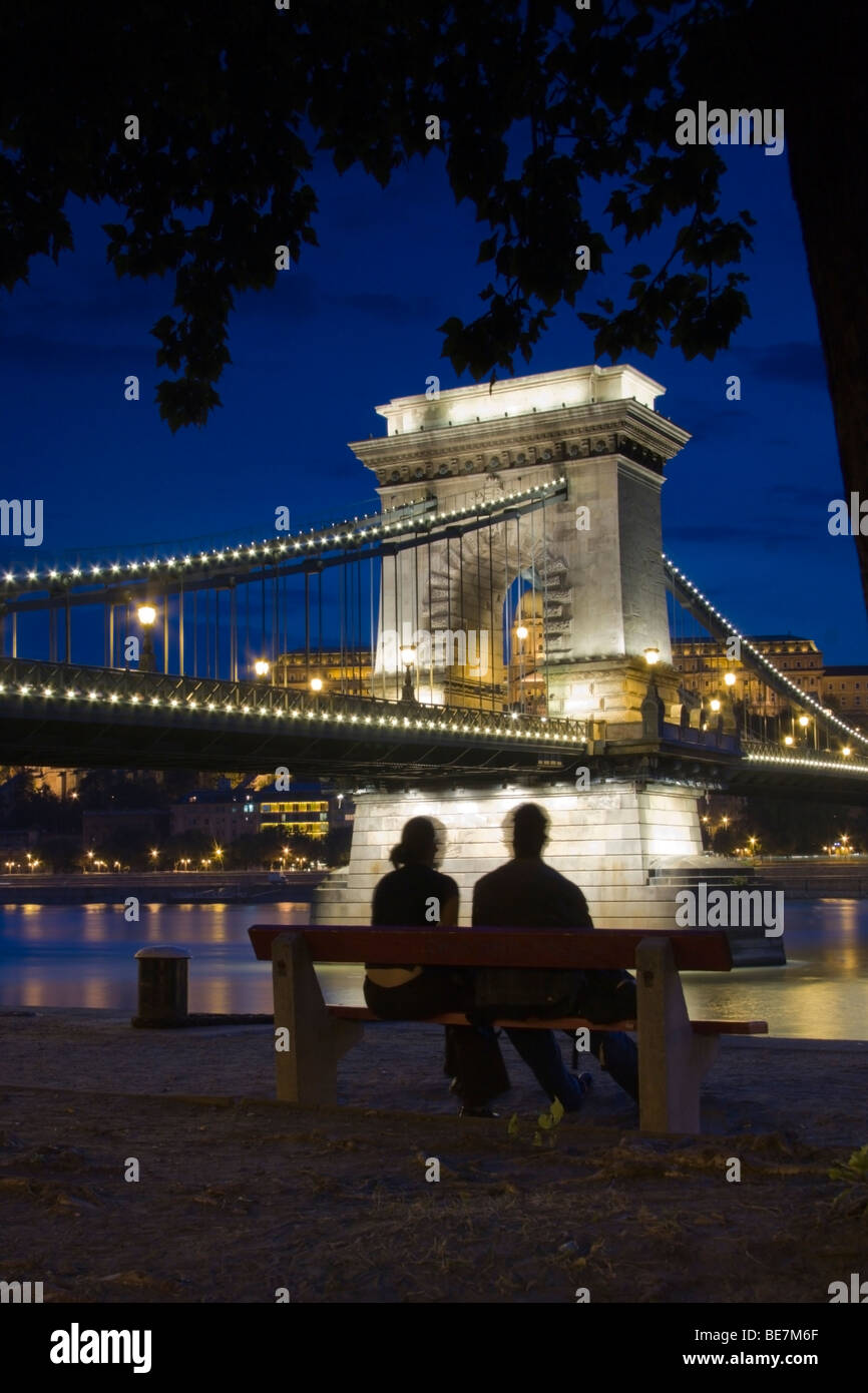 A couple sit on a bench in front of Budapest's famous Chain Bridge, illuminated in the early evening. Stock Photo