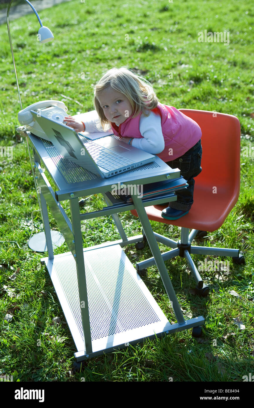 Little girl standing on office chair by desk in field, playing with laptop computer, looking at camera Stock Photo