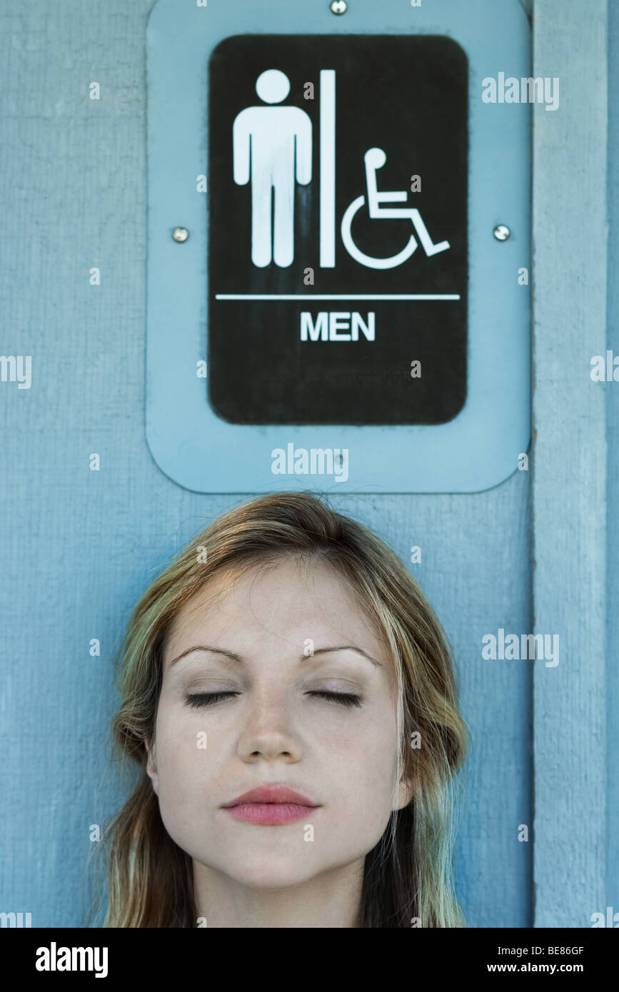 Woman in front of men's restroom sign, eyes closed Stock Photo