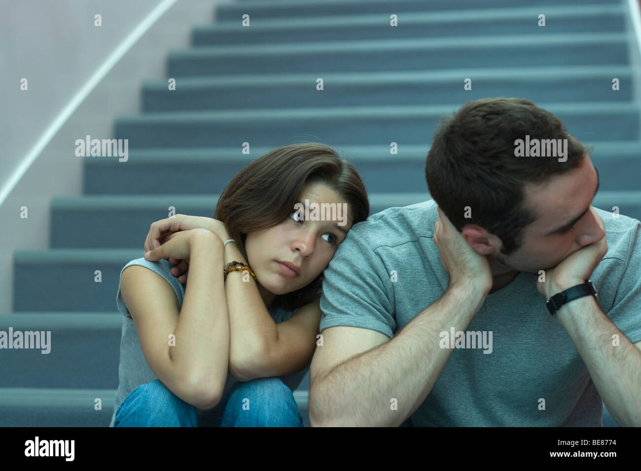 Young couple sitting side by side on stairs, woman leaning head on man's shoulder, man looking away Stock Photo