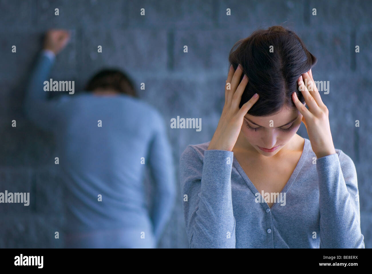 Young woman holding head, man in background with back turned leaning against wall Stock Photo