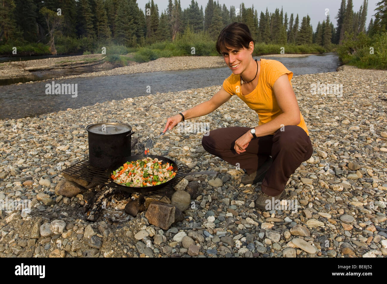 Young woman cooking, frying chicken stir fry on a camp fire, pot, pan, spatula, gravel bar, upper Liard River, Yukon Territory, Stock Photo