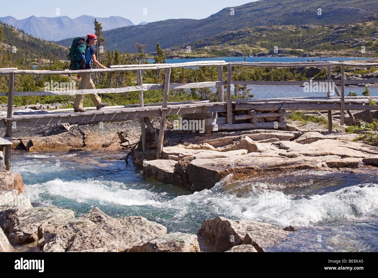 Young woman crossing creek, wooden bridge, Deep Lake behind, hiking, backpacking, hiker with backpack, historic Chilkoot Trail, Stock Photo