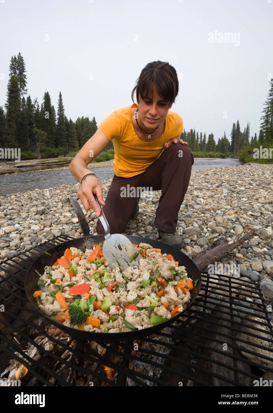 Young woman cooking, frying chicken stir fry on a camp fire, pan, spatula, gravel bar, upper Liard River, Yukon Territory, Cana Stock Photo
