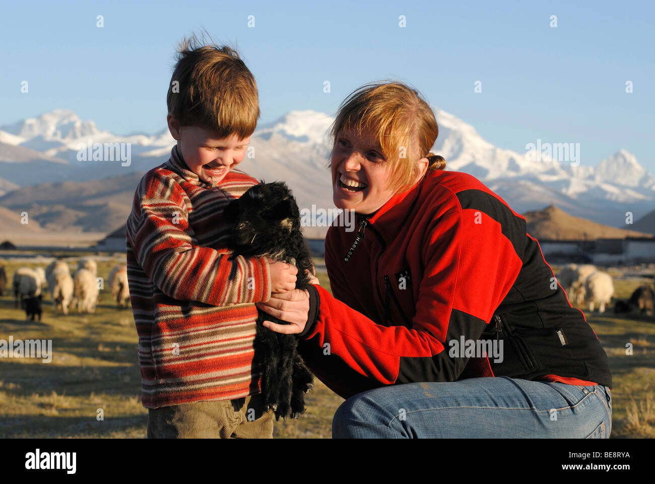 Little girl joyfully holding a little black lamb, alongside a smiling woman in front of the snowy mountains of Cho Oyo, 8112 m, Stock Photo