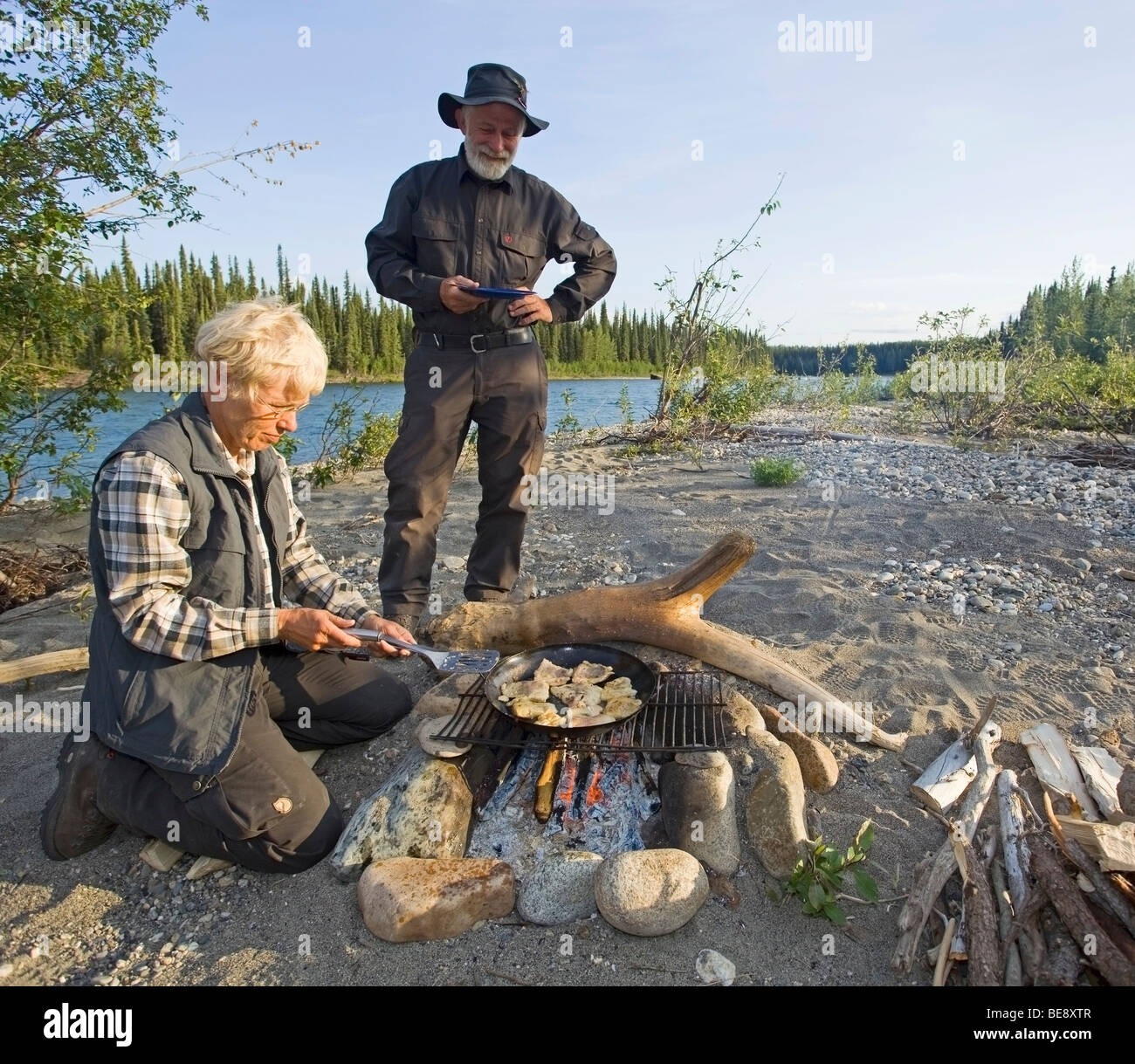 Woman cooking, frying fish fillets in a pan on a camp fire, man with plate waiting behind, upper Liard River, Yukon Territory,  Stock Photo