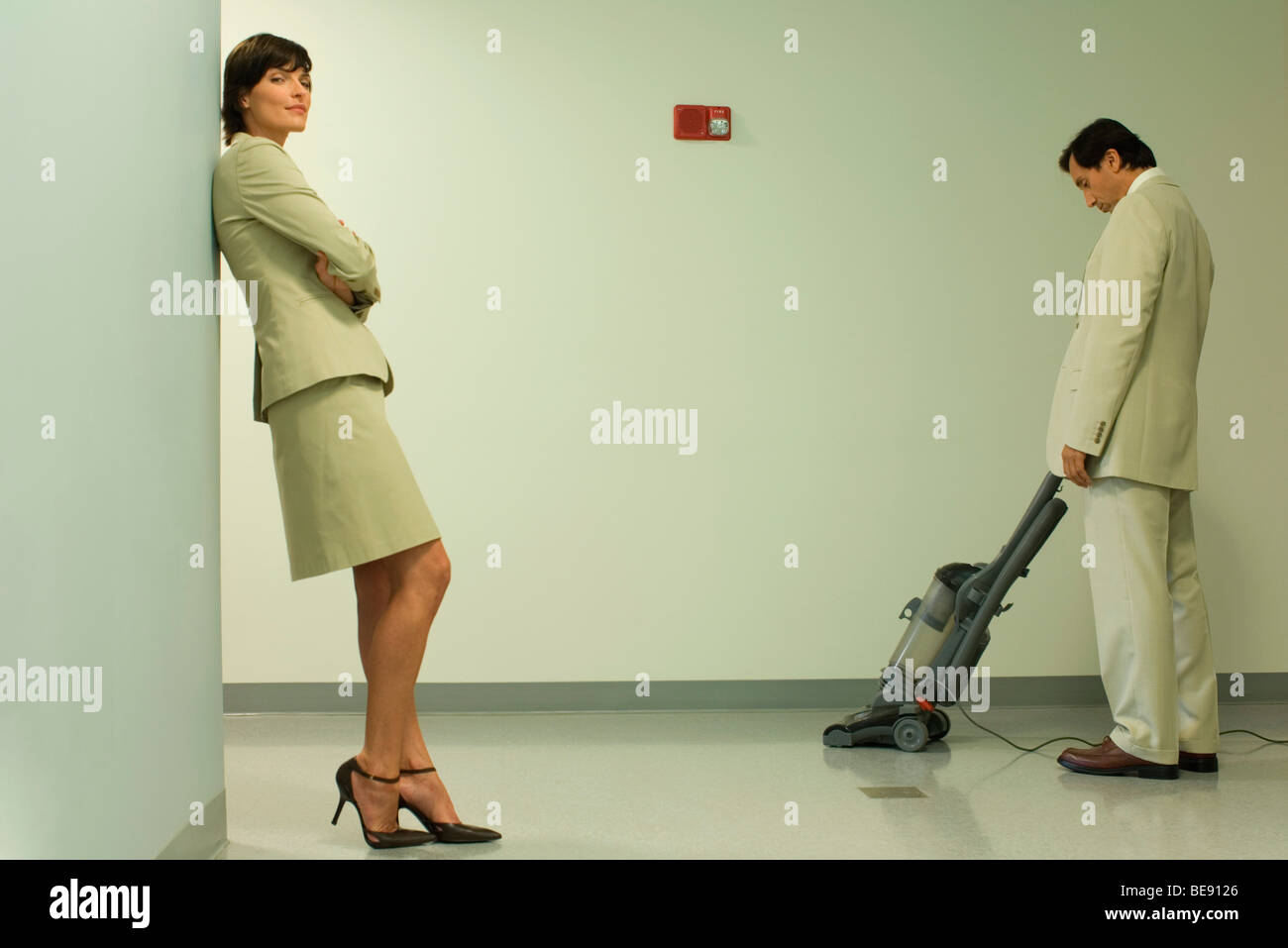 Well-dressed woman leaning against wall with arms folded, man vacuuming in background Stock Photo