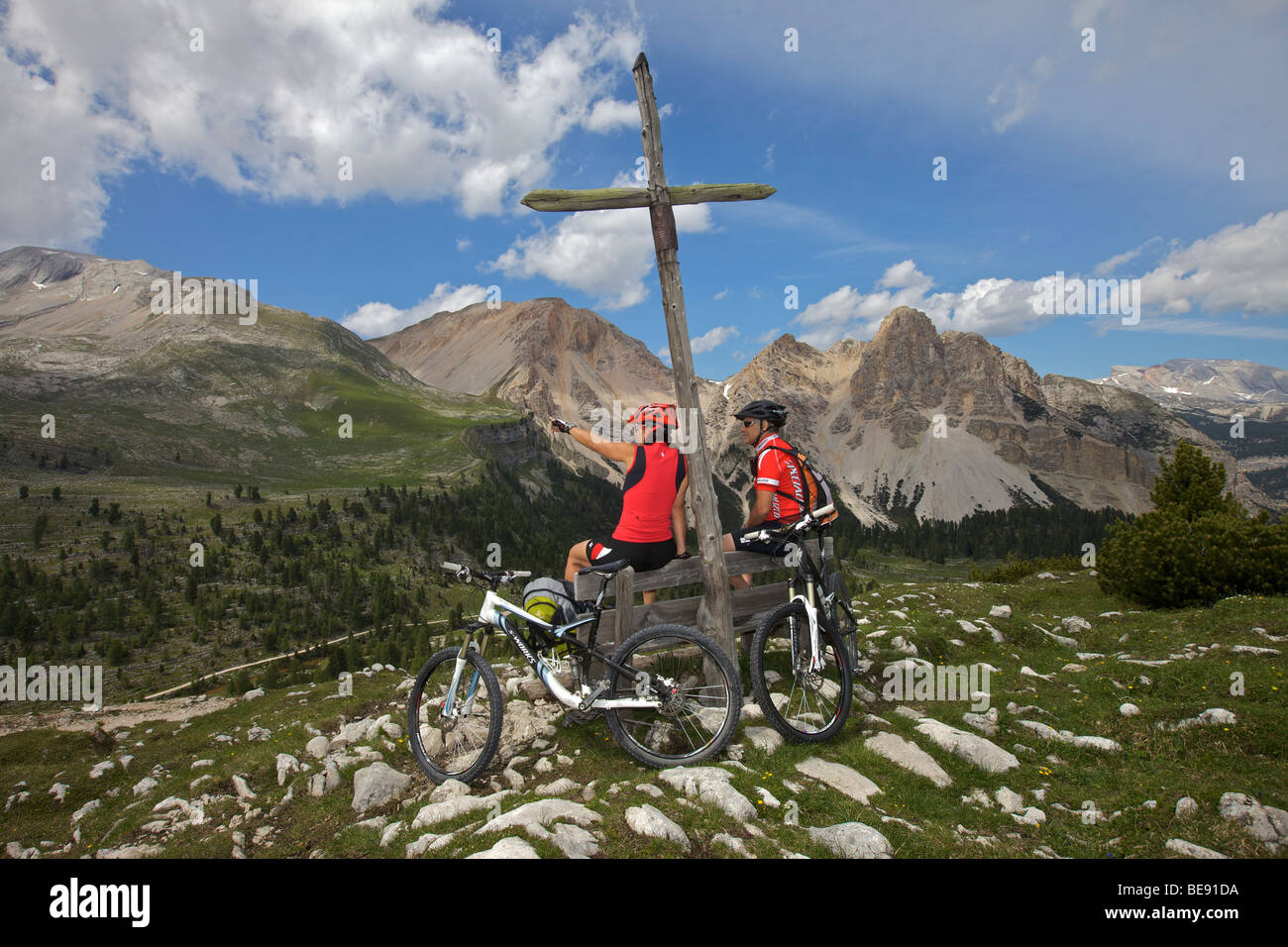 Mountain bike riders having a rest stop on a wooden bench in front of a wooden cross on the Limo Pass in Fanes-Sennes-Prags Nat Stock Photo