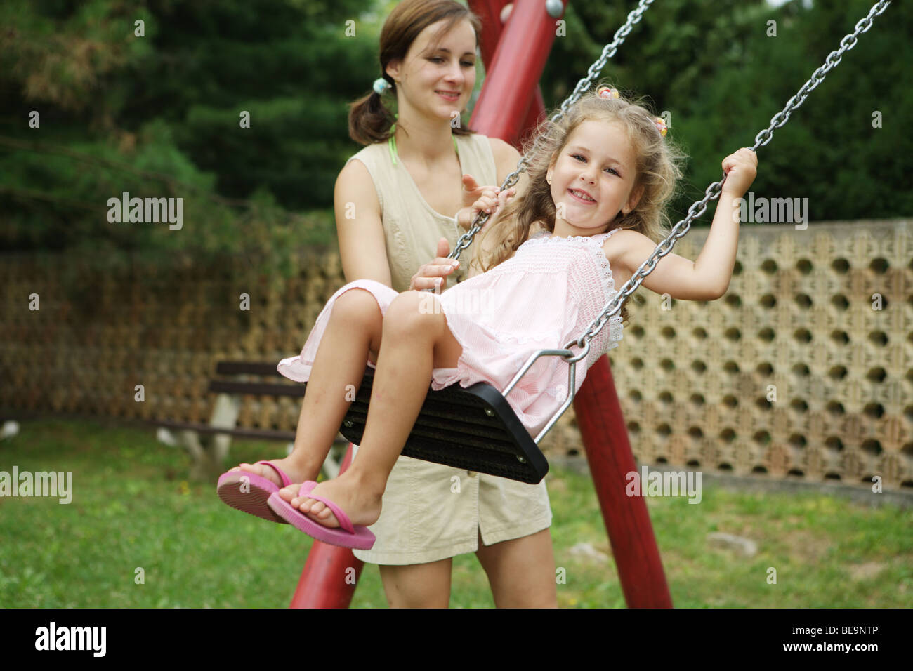 Mother and daughter playing in playground. Child is swinging on swing. Stock Photo