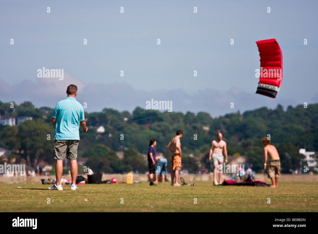 A man relaxing flying a red sparless kite at Pool Quay park on a sunny summers day. Other people relax in the background. Stock Photo